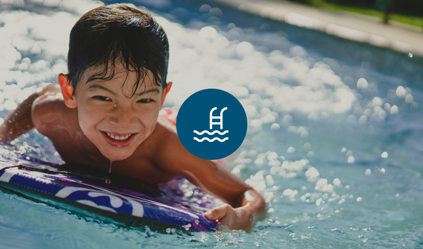 Photograph of a young boy in a pool, smiling and floating on a foam kickboard.